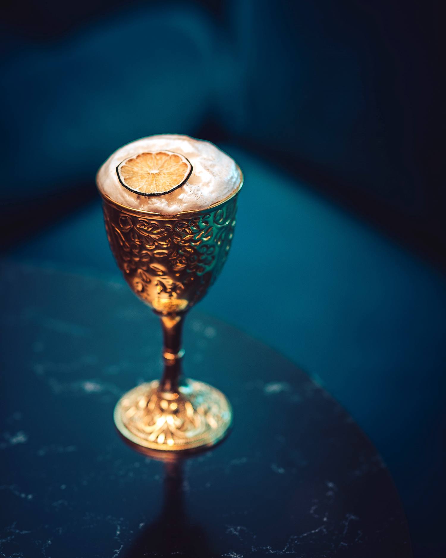 Gold ornate goblet with a creamy beverage topped with a lime slice, placed on a dark marble table against a dark background.