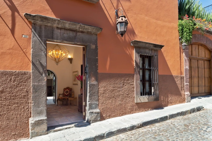 Rustic colonial entrance with an open door, revealing an elegant interior with a chandelier and vintage chair.