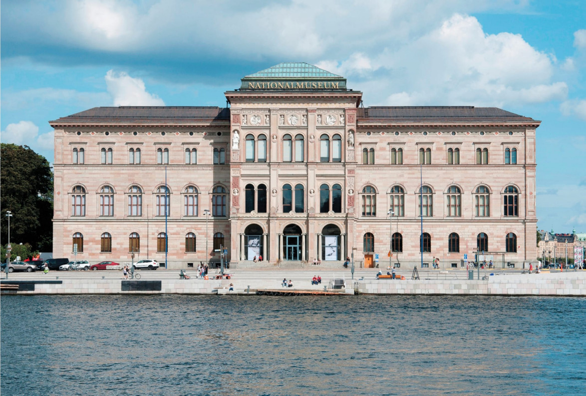 Front view of a large, historic building with arches and decorative features, set against a blue sky and water in the foreground.