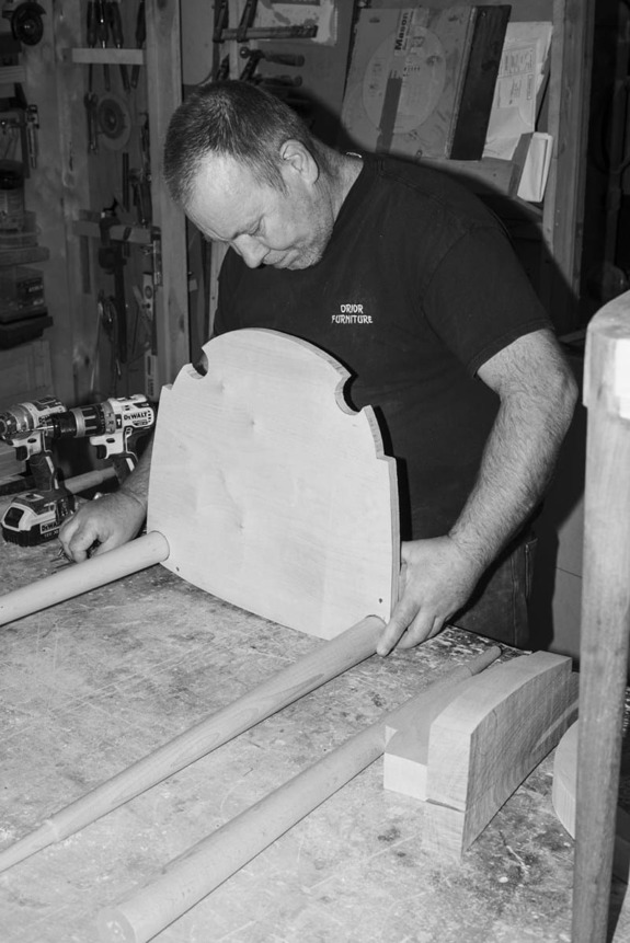 Woodworker assembling a chair backrest in a workshop, surrounded by tools and wooden components.