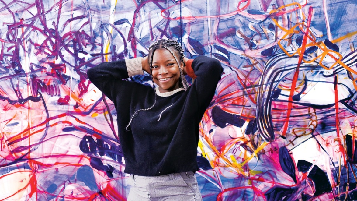 Smiling person standing in front of a colorful abstract mural, wearing a black sweater and gray pants.