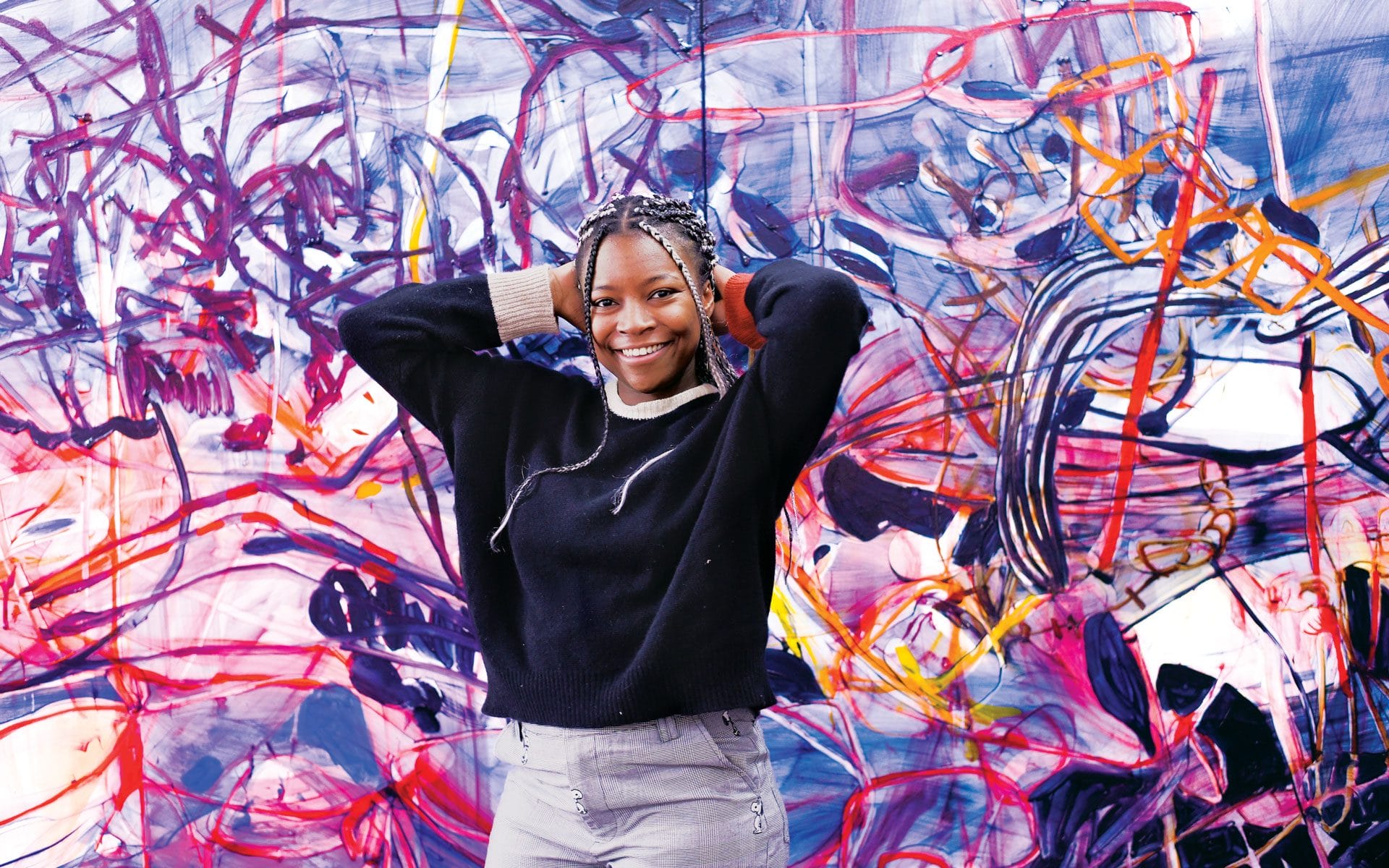 Smiling person standing in front of a colorful abstract mural, wearing a black sweater and gray pants.