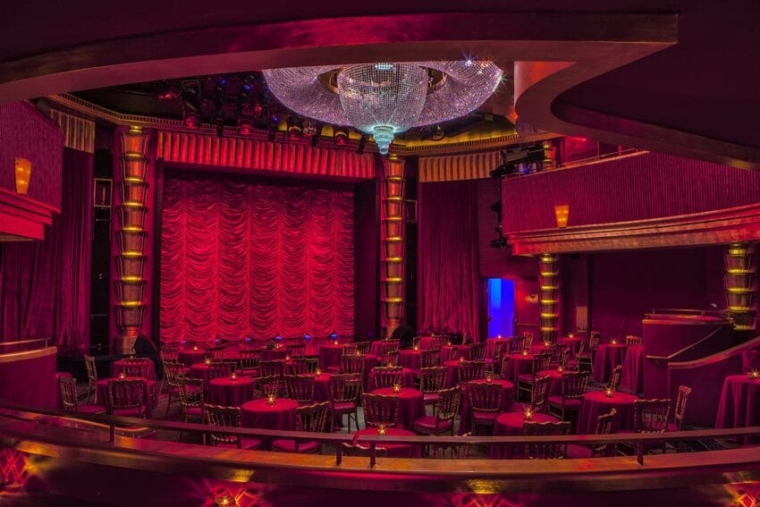 Elegant theater with red curtains, round tables, chandeliers, and stage lighting, creating a warm and inviting atmosphere.