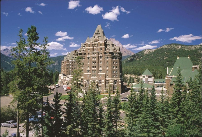 Majestic castle-like hotel surrounded by lush pine trees with a mountain backdrop under a clear blue sky.