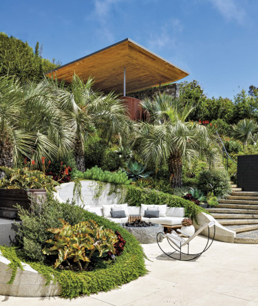 Modern patio with lounge chairs surrounded by lush greenery, palm trees, and a contemporary wooden roof structure.