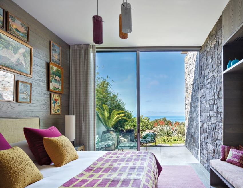 Modern bedroom with colorful decor, large glass doors opening to a garden with ocean view, framed art on walls.