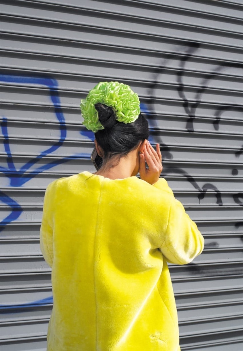 Person with green hair accessory in yellow coat stands in front of a graffiti-covered metal wall.
