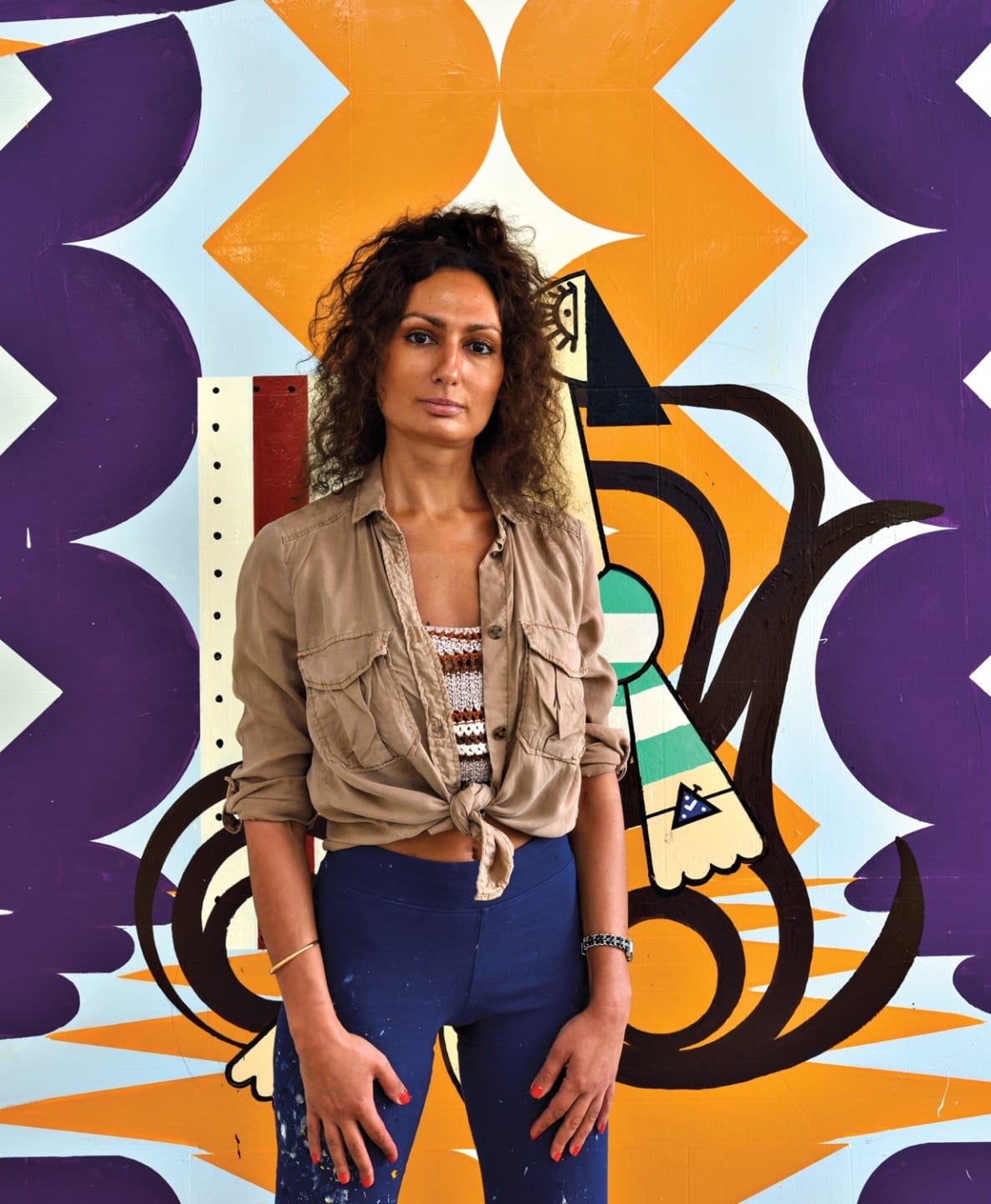 Person standing in front of colorful geometric wall art, wearing a beige shirt and blue pants, with a thoughtful expression.