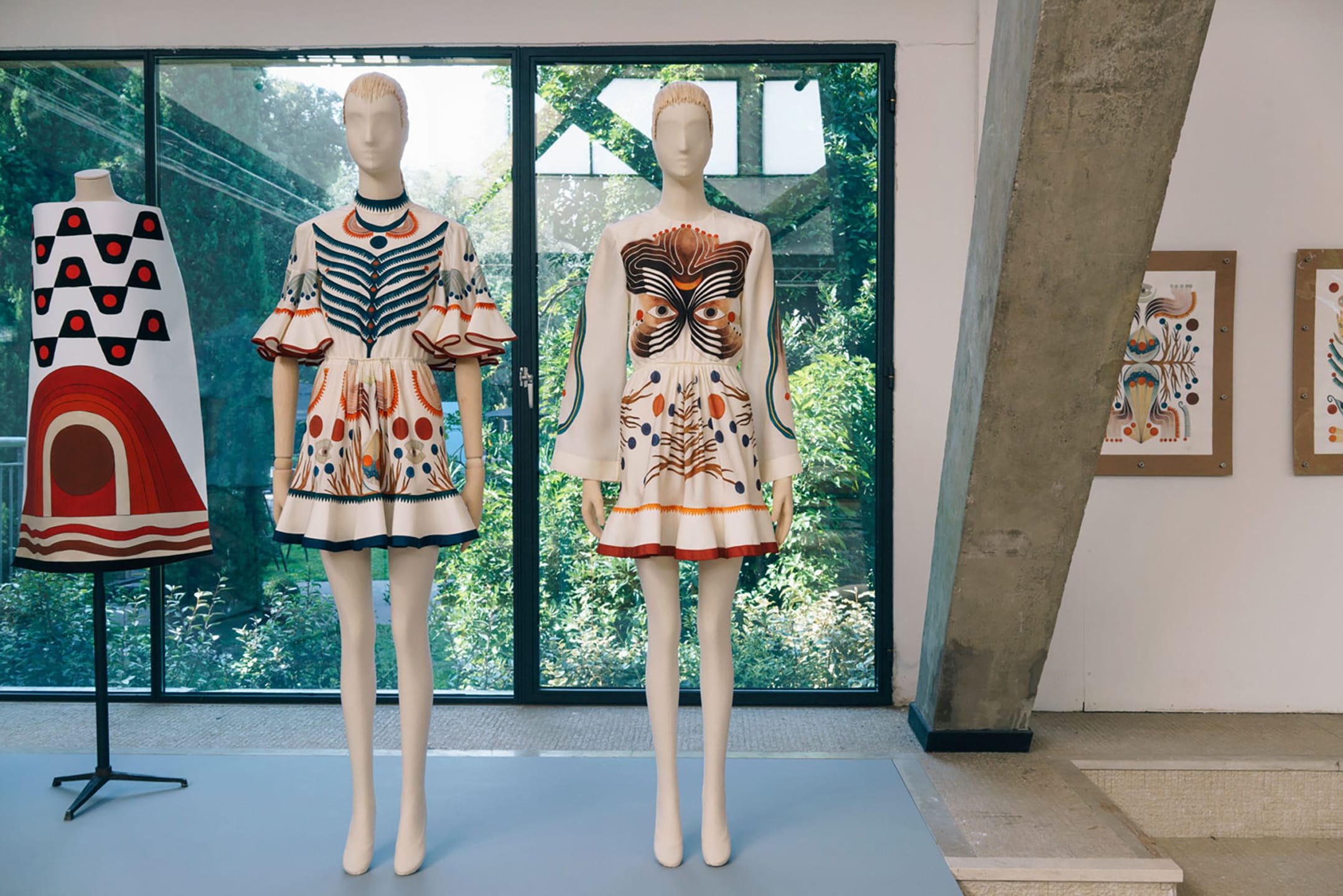 Two mannequins wear colorful abstract patterned dresses in a modern gallery with large windows and artwork on walls.