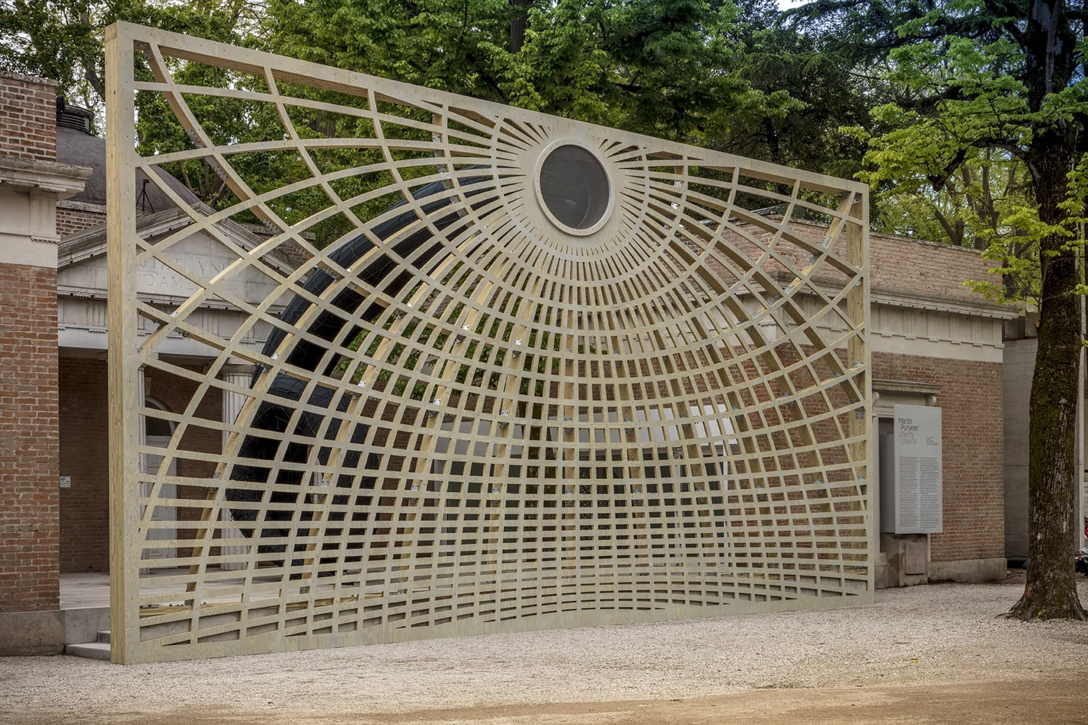 Outdoor art installation featuring a large wooden grid structure with a circular cutout set against a background of trees.