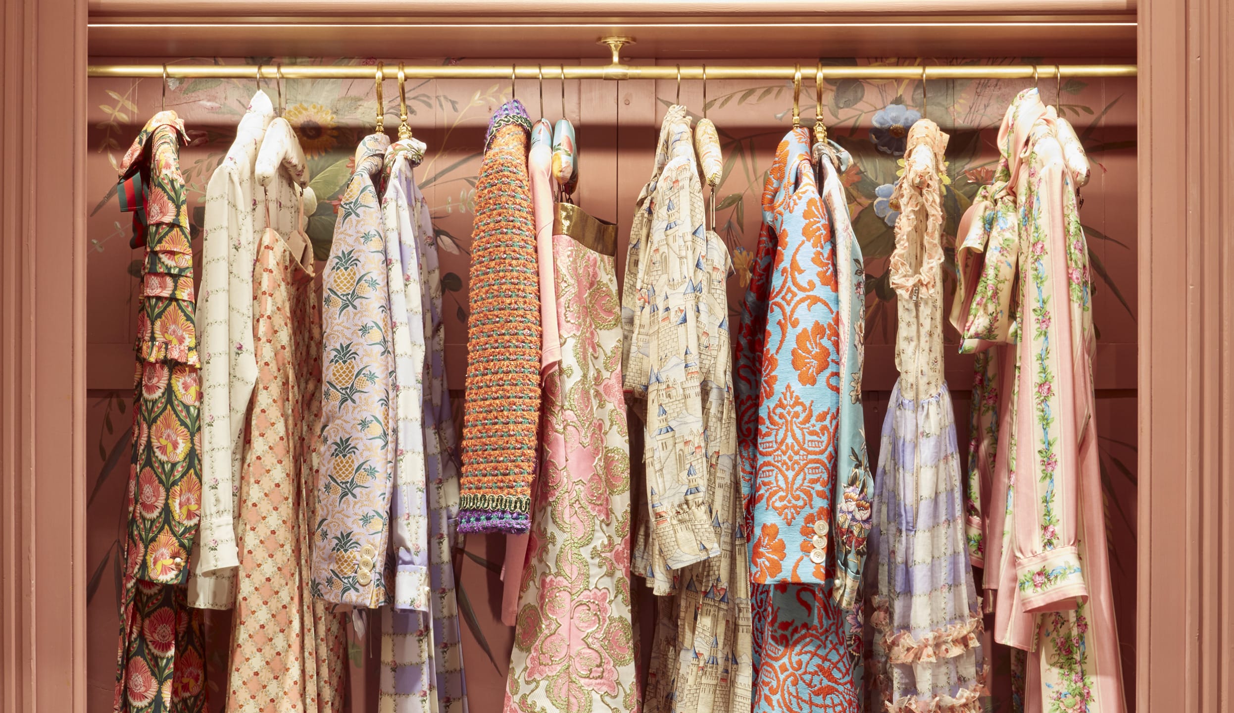 Colorful patterned vintage dresses hanging on a gold clothing rack against a decorative floral wall.
