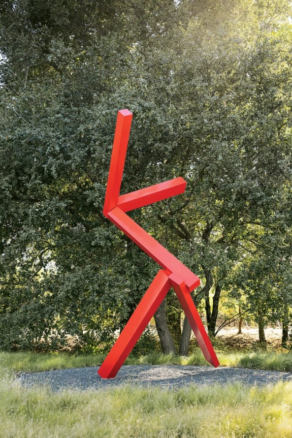 Red abstract sculpture of a dancing figure in front of green trees.