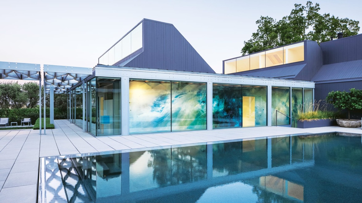 Modern house with glass walls, blue accents, and a large patio next to a reflective swimming pool at dusk.