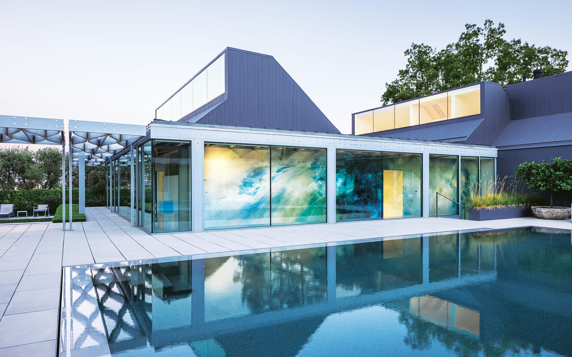 Modern house with glass walls, blue accents, and a large patio next to a reflective swimming pool at dusk.