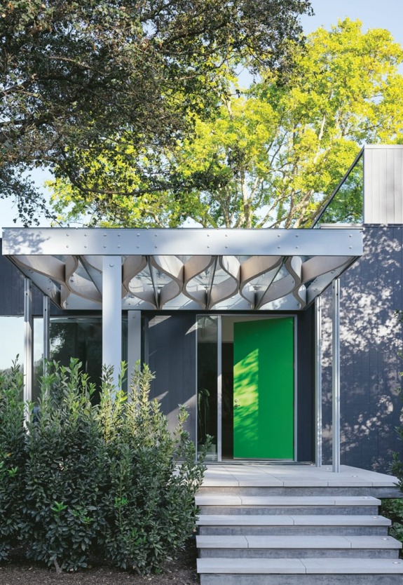 Modern building entrance with geometric canopy, green door, and lush foliage against a backdrop of trees and clear sky.