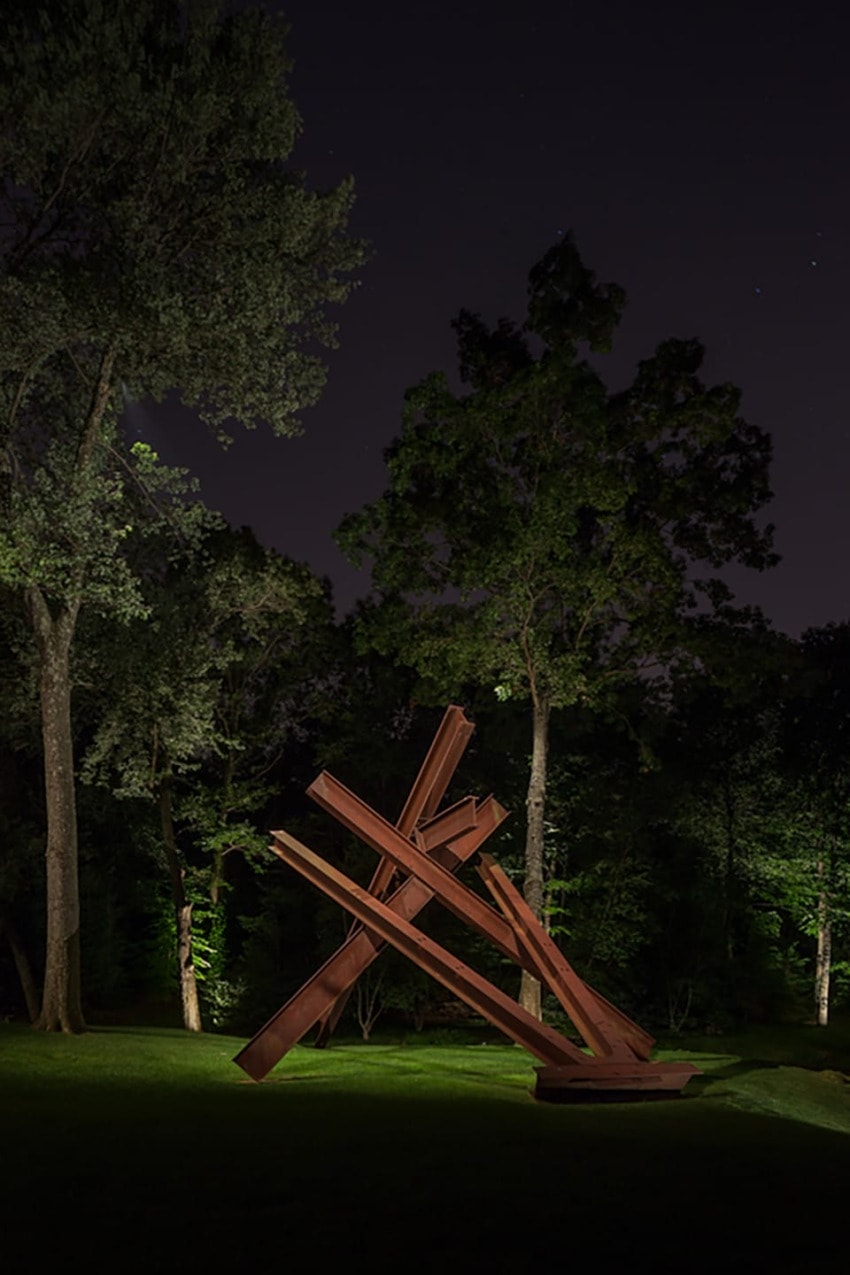 Steel beam sculpture in a forest setting at night, with tall trees and a dark sky in the background.