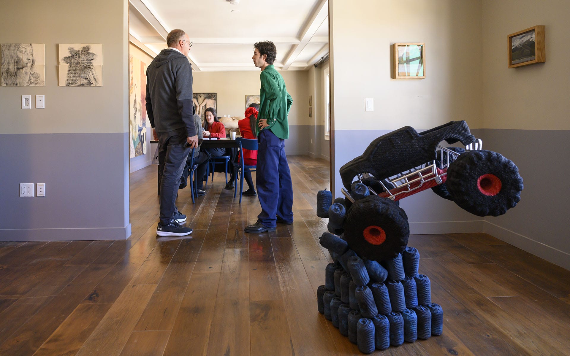 People conversing in a modern art gallery with a sculpture of a monster truck made from dark materials in the foreground.