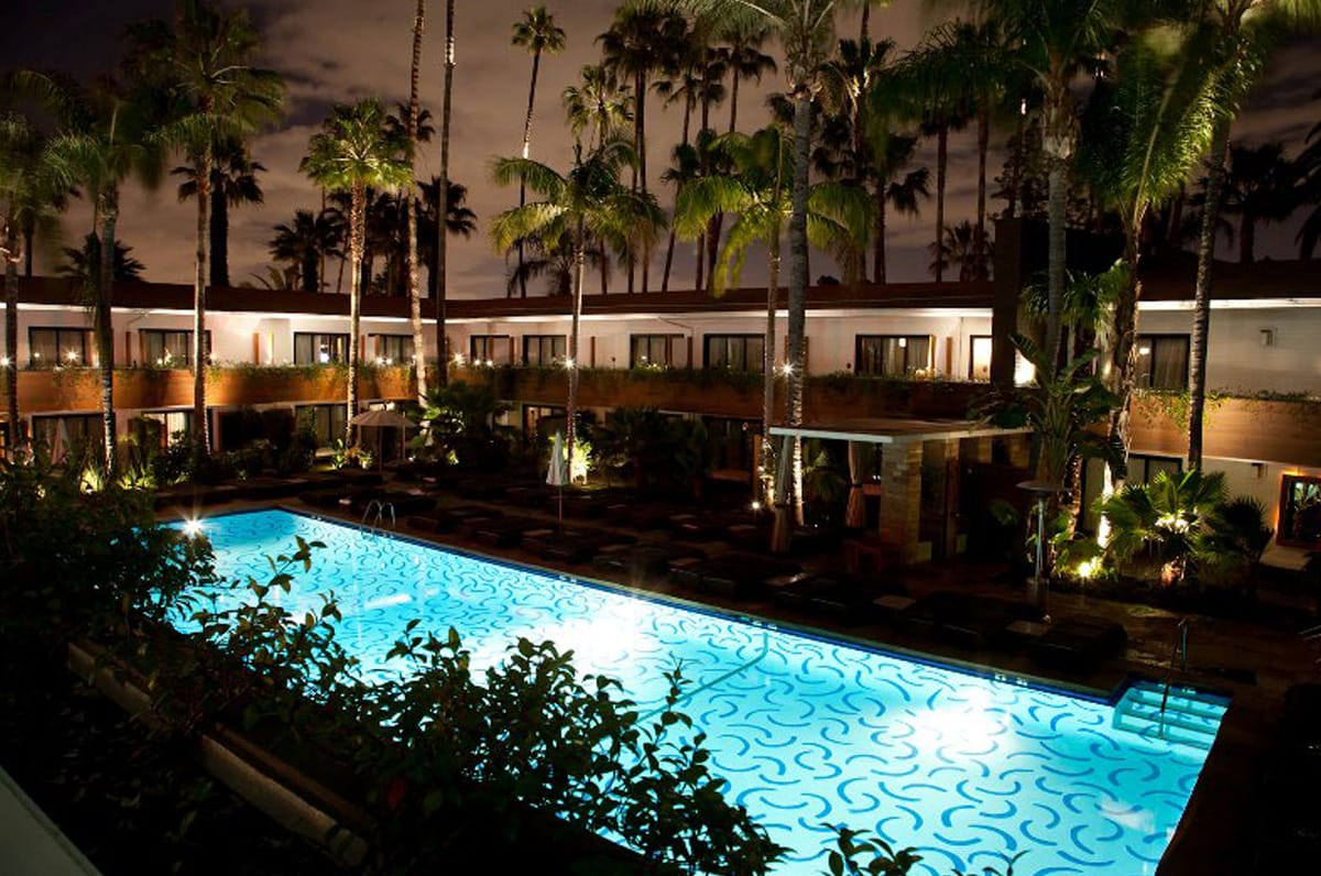 Illuminated outdoor pool surrounded by palm trees and hotel buildings at night.