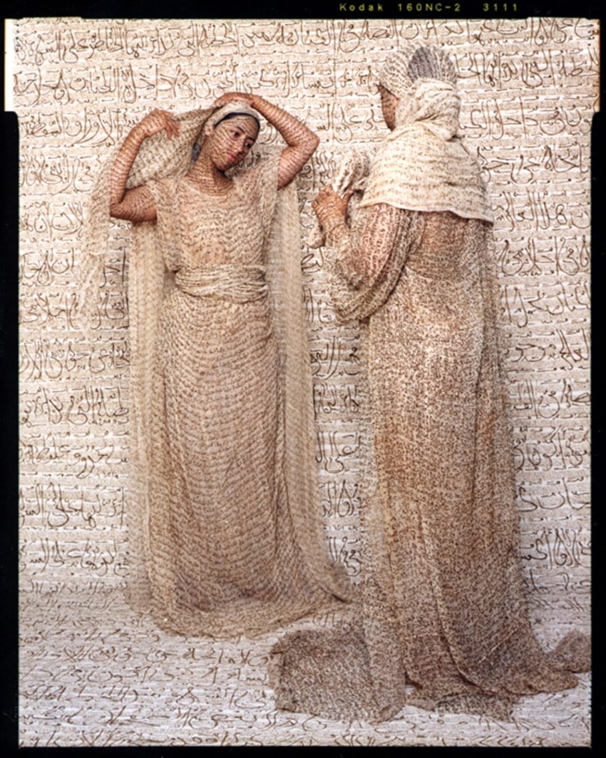 Two women in beige attire stand against a background of intricate Arabic calligraphy.