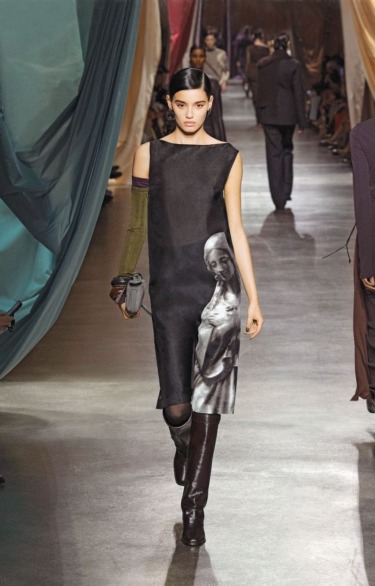Model walking on a runway wearing a black dress with a classical art print, surrounded by draped fabric and audience.