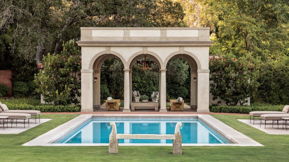 Outdoor pool with a stone pergola and lush green surroundings, featuring lounge chairs and a reflective water surface.