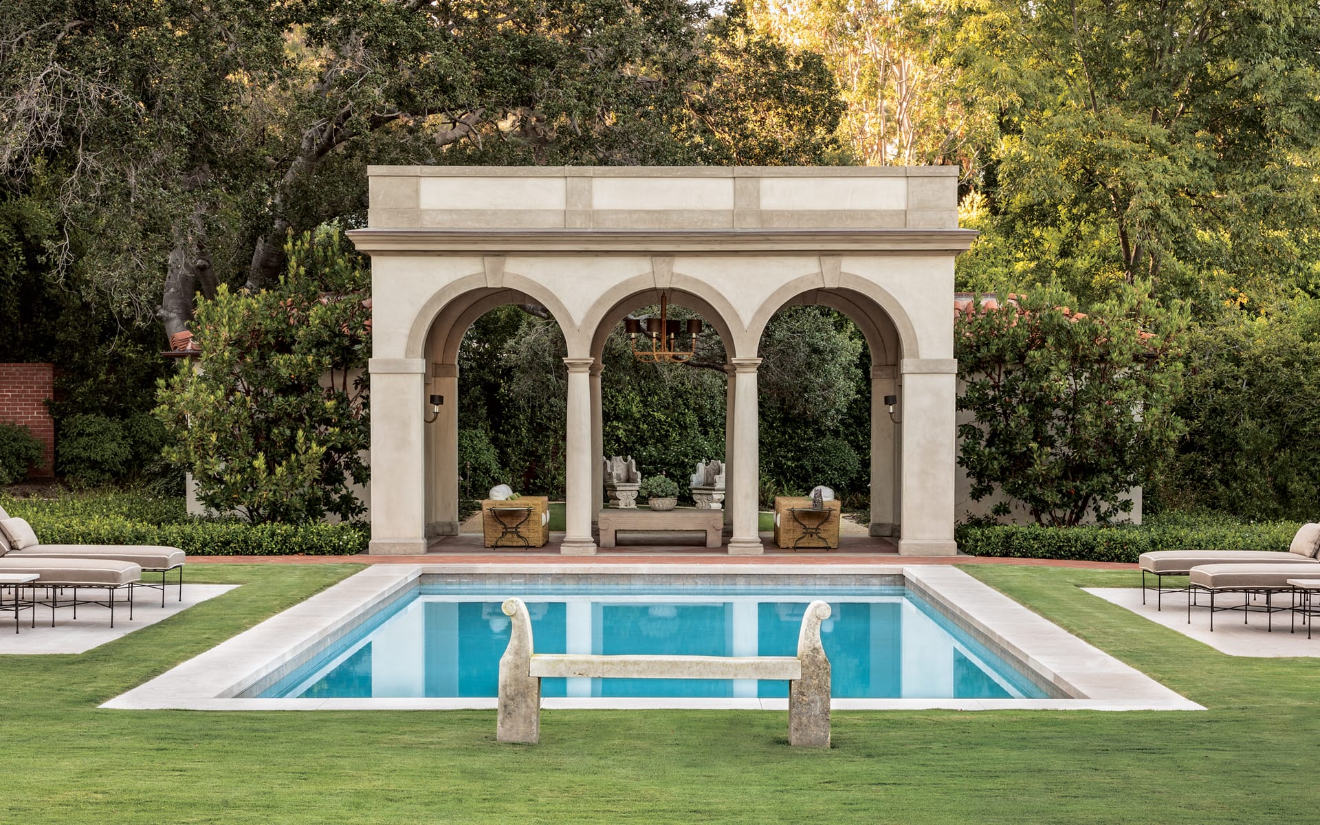Outdoor pool with a stone pergola and lush green surroundings, featuring lounge chairs and a reflective water surface.