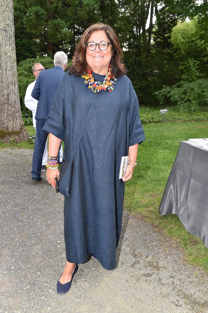 A person wearing a blue dress and colorful necklace standing outdoors on a gravel path with trees in the background.