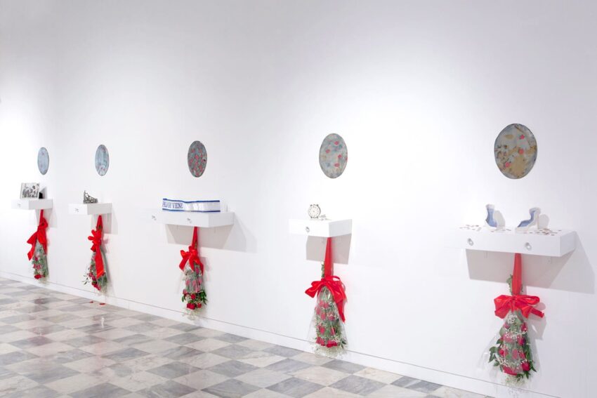Gallery wall display with decorative plates and bouquets beneath shelves, each holding art pieces, on a tiled floor.