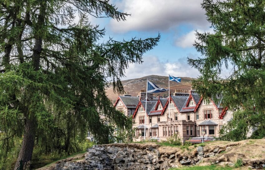 Historic stone mansion surrounded by lush trees with Scottish flag waving on the roof, mountains visible in background