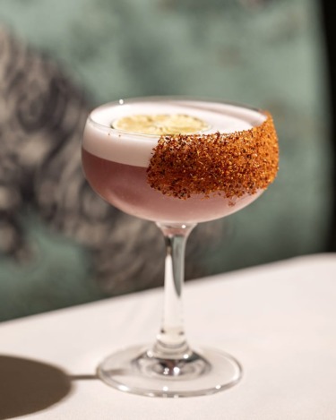 Cocktail in a stemmed glass with a frothy top, garnished with a lime slice and chili rim, on a white tablecloth.