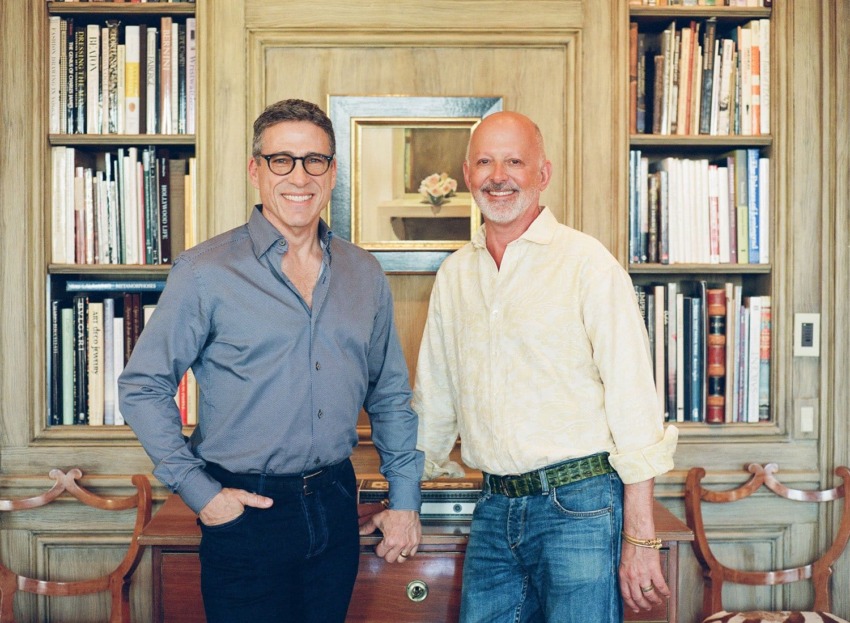 Two men smiling and standing in front of a wooden bookshelf filled with books in a cozy room.