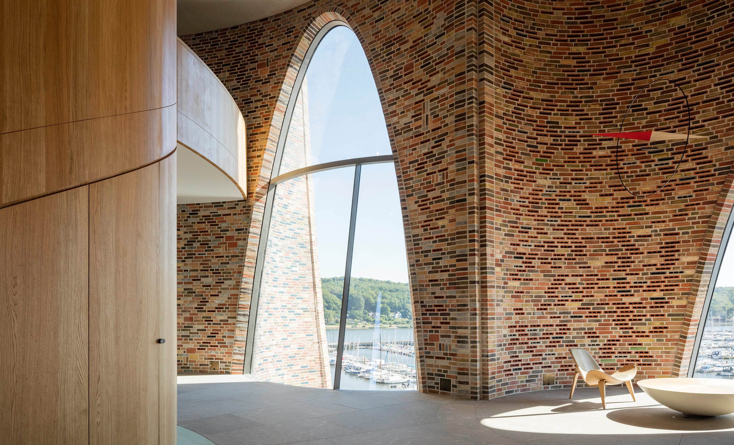 Modern interior with large arched windows, brick walls, a wooden chair, and scenic view of marina outside.