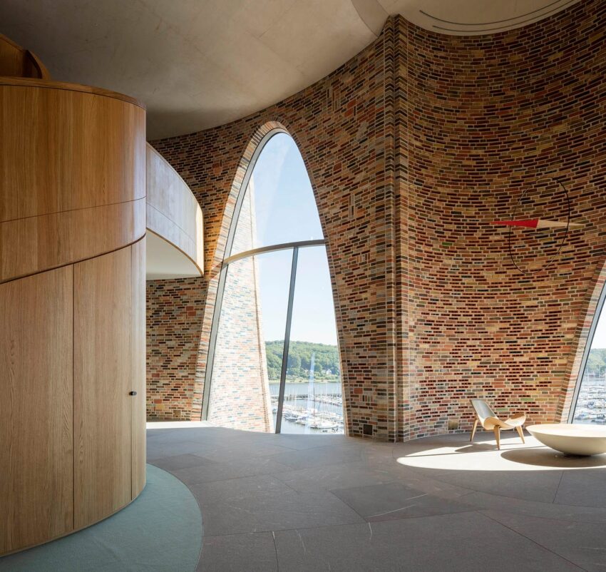 Modern interior with brick walls, large curved windows, and minimalist chairs overlooking a scenic outdoor view