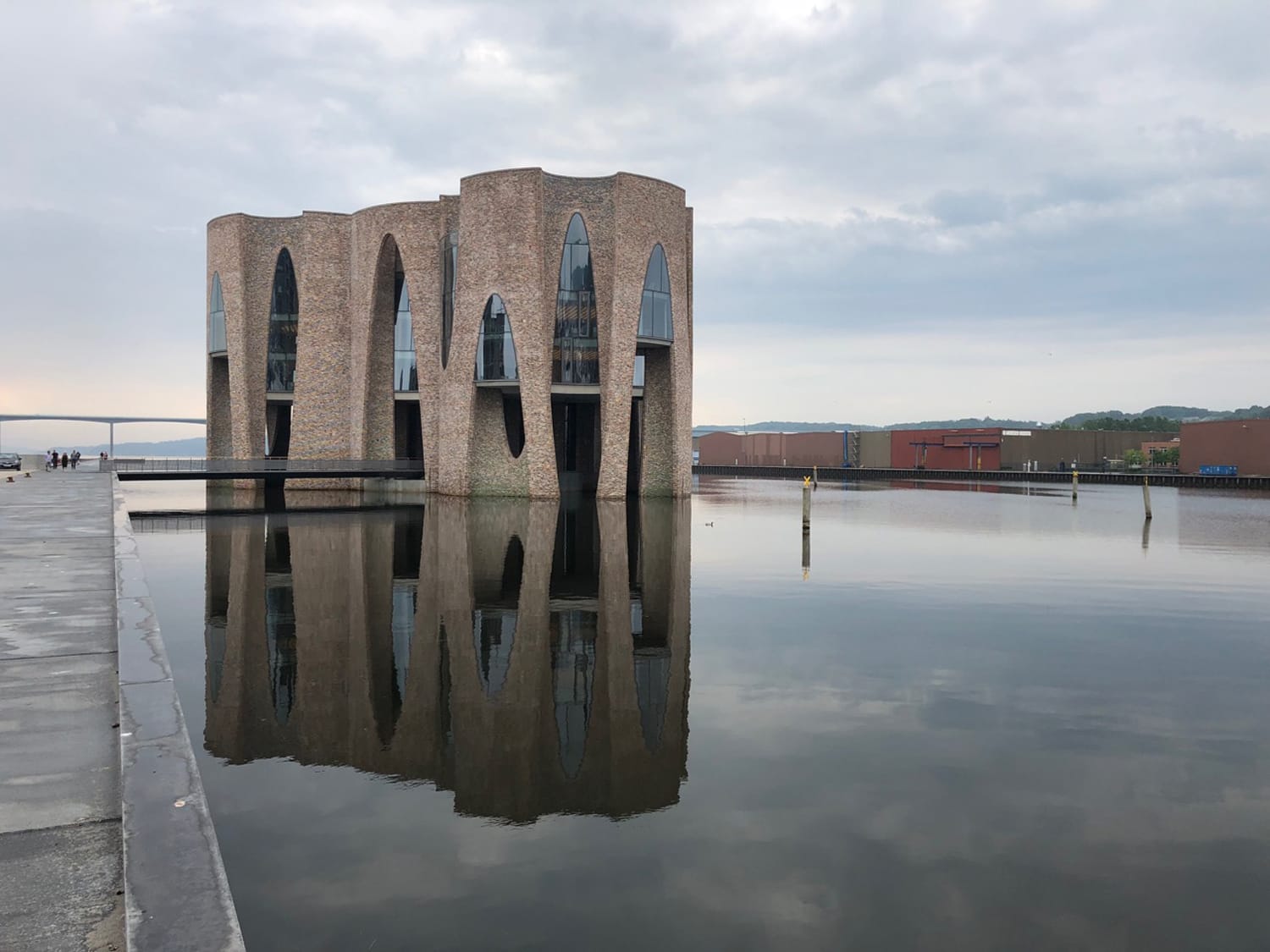 Unique brick building with large arches reflected in calm waters under a cloudy sky.