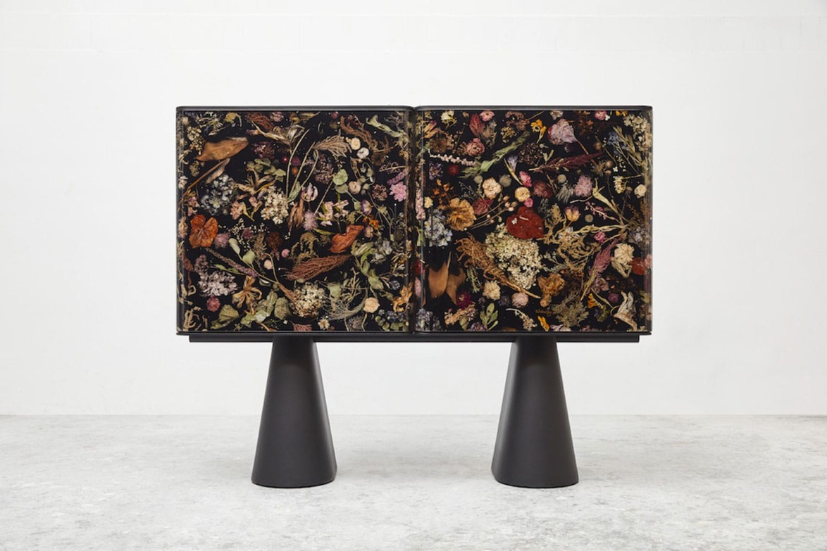 Decorative cabinet with floral and bird design, standing on two black cylindrical legs, placed in a minimalist space.