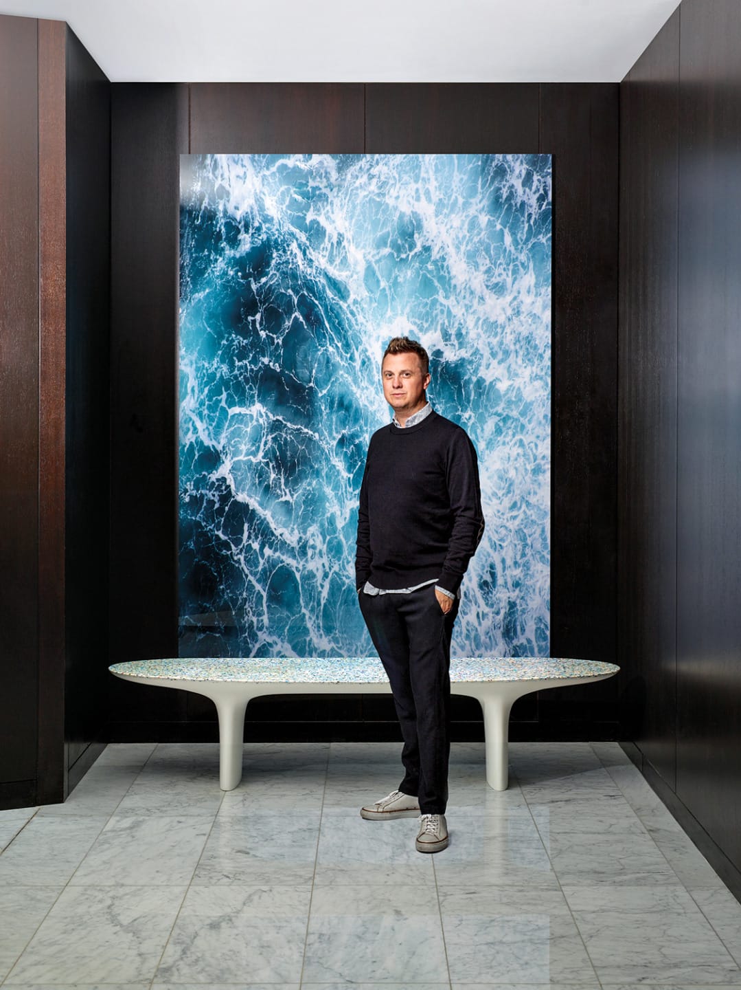 Man standing in front of large ocean-themed artwork, wearing dark clothing and grey shoes, in a modern room with marble floors.