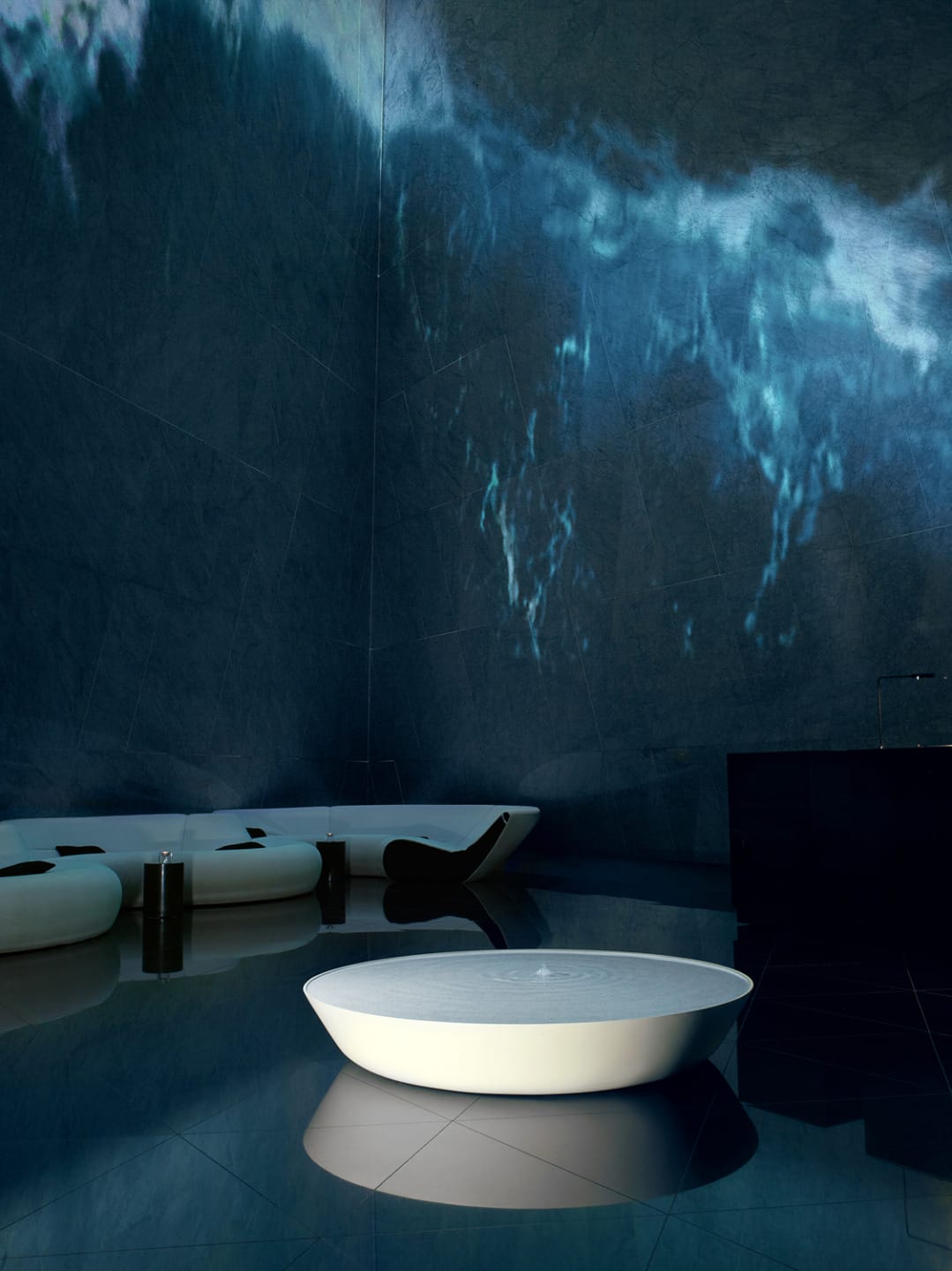 Futuristic lounge with sleek white furniture, dark floors, and a digital wave projection on a tall wall.