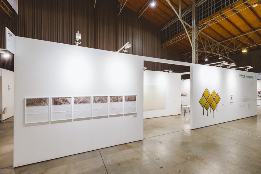 Art exhibition with framed pictures on white walls in a spacious gallery with high ceilings and industrial lighting.