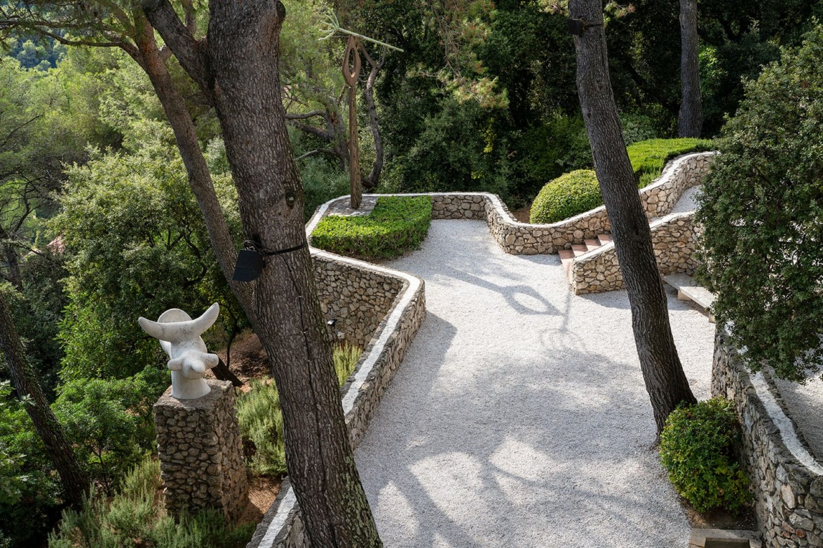 Stone pathway in a garden with trees and a sculpture, surrounded by lush greenery and stone walls.