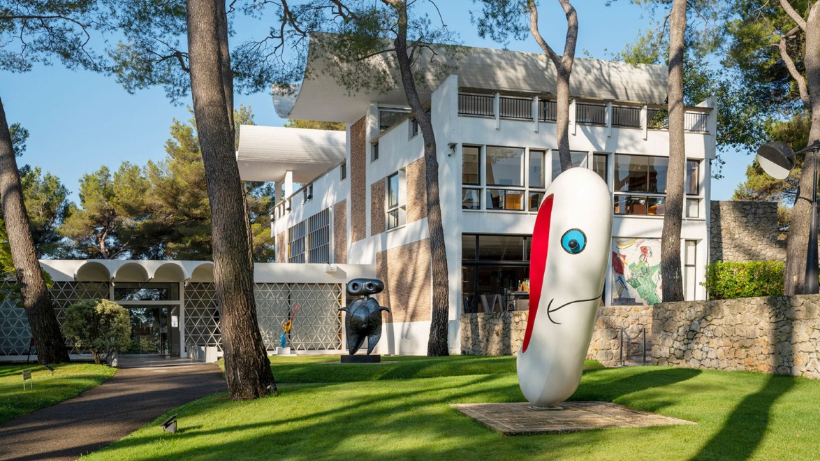 Modern art sculptures on a lawn in front of a contemporary white building surrounded by trees.