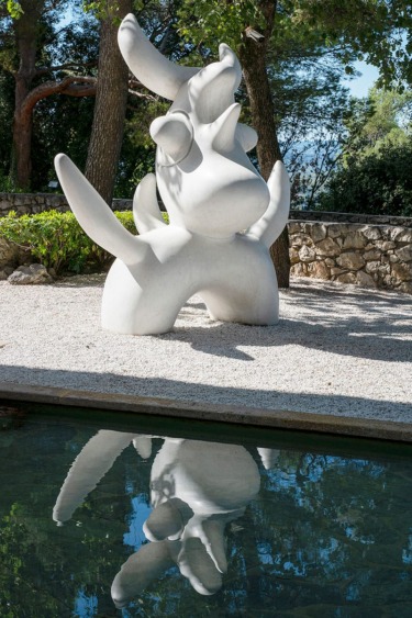 Abstract white sculpture of a creature with horns, reflecting in a pond, set in a wooded garden under a clear blue sky.