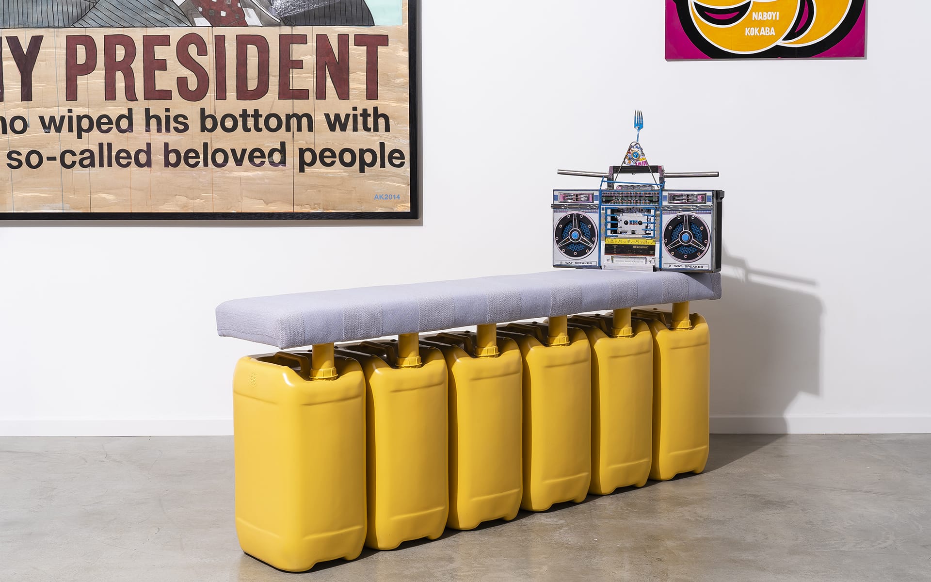 A bench made of yellow jerry cans with a stereo on top in an art gallery setting with various wall art.