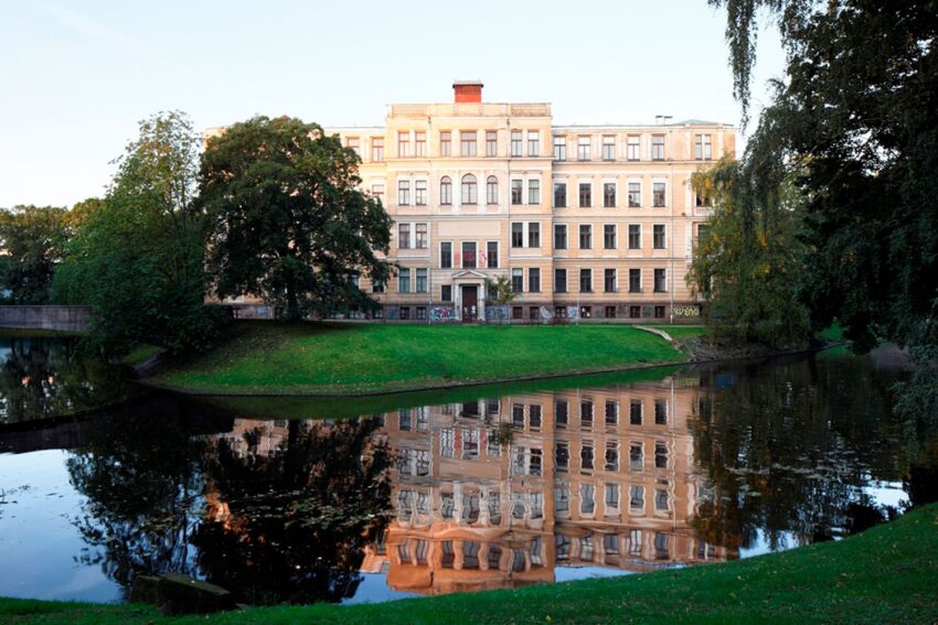 Historic building reflected in a calm pond surrounded by green grass and trees under a clear blue sky.