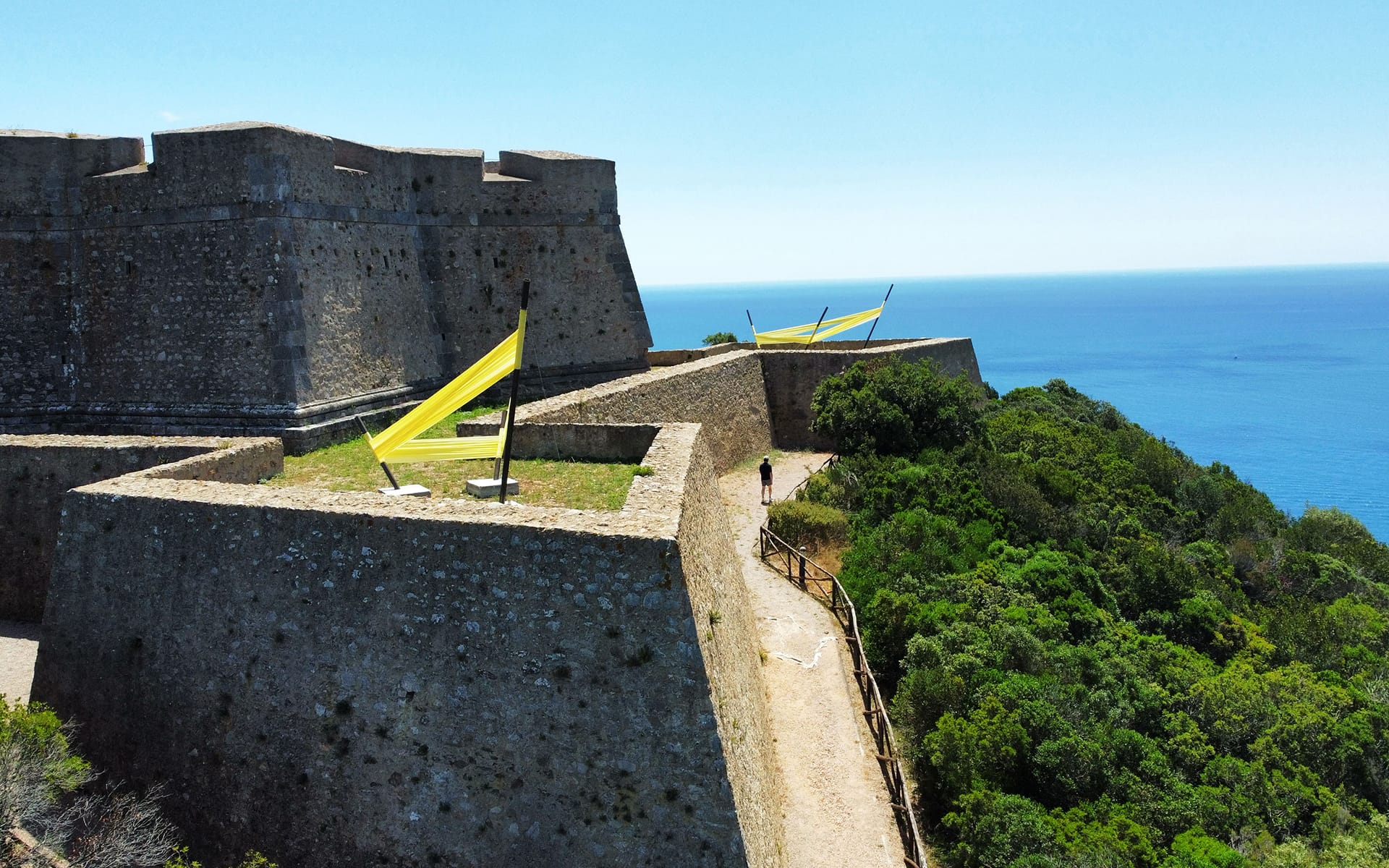 Historic stone fortress with yellow flags, surrounded by greenery, overlooking the sea on a clear day.