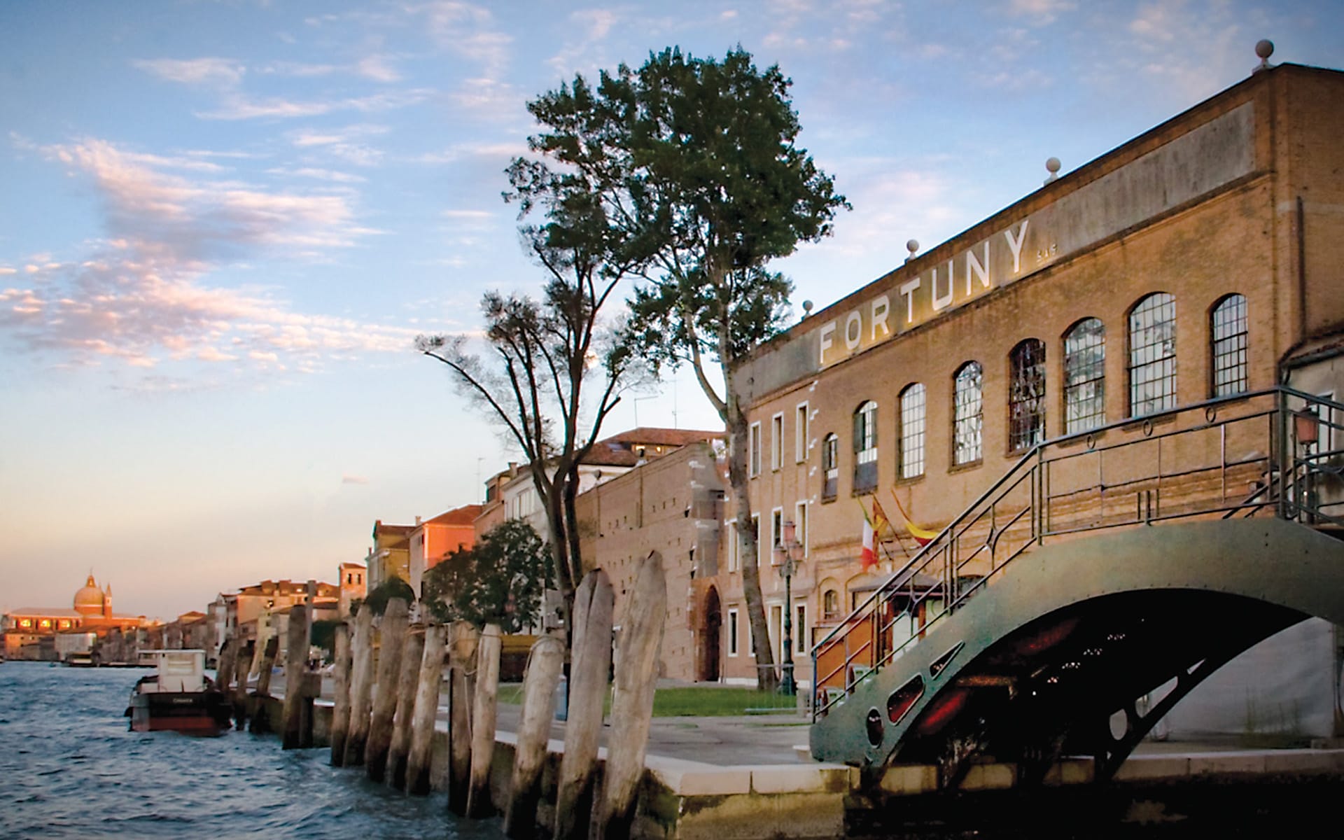 View of a canal in Venice at sunset with historic brick buildings, a tree, and a bridge over the water.