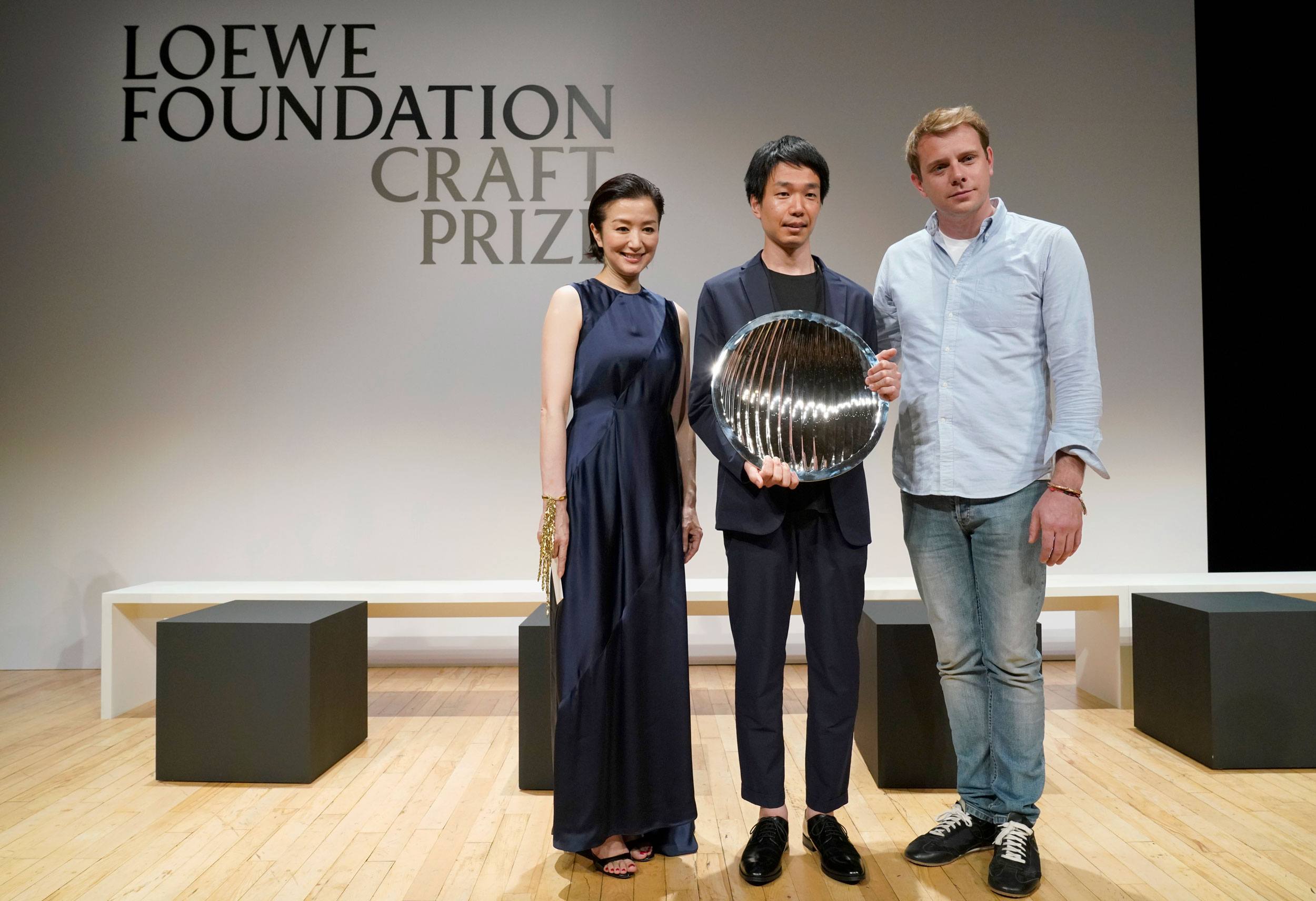 Three individuals standing on a stage, with one holding a reflective circular award, under a sign saying Loewe Foundation Craft Prize.