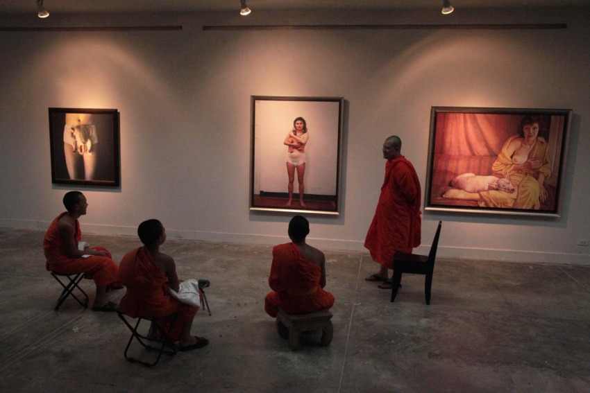 Monks in orange robes seated on stools, observing artwork at a gallery with dim lighting.