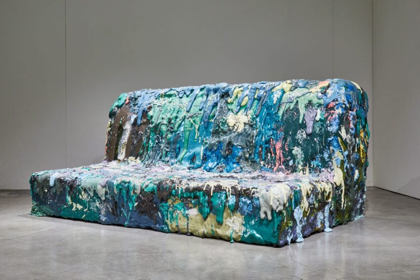 Abstract sculpture resembling a melting couch with vibrant turquoise, blue, and multicolored layers on a concrete floor.