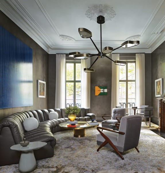 Modern living room with a large sectional sofa, stylish chandelier, abstract art, and bright natural light from tall windows.