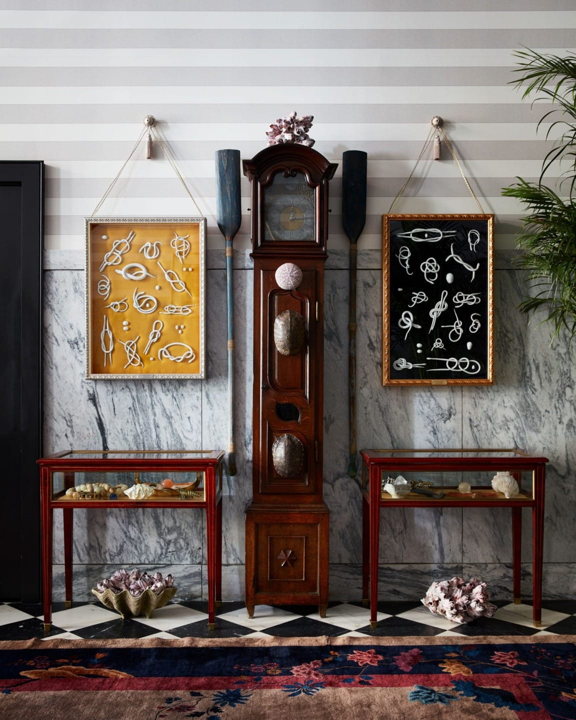 Grandfather clock between two framed knot displays, with wooden tables and decorative items below, against a striped wall.